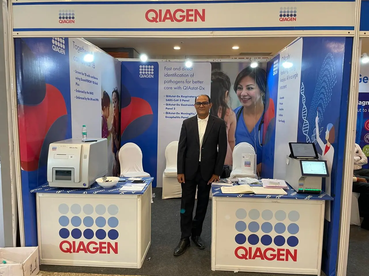 A man stands in a QIAGEN branded exhibition booth displaying diagnostic instruments.