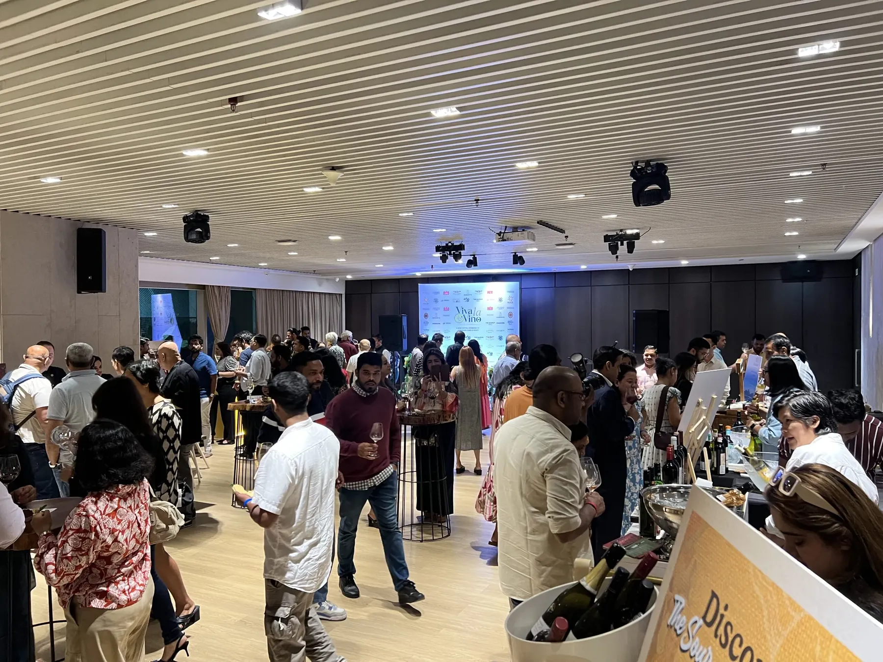 Guests mingle and taste wine at the Viva La Vino festival before a branded backdrop.