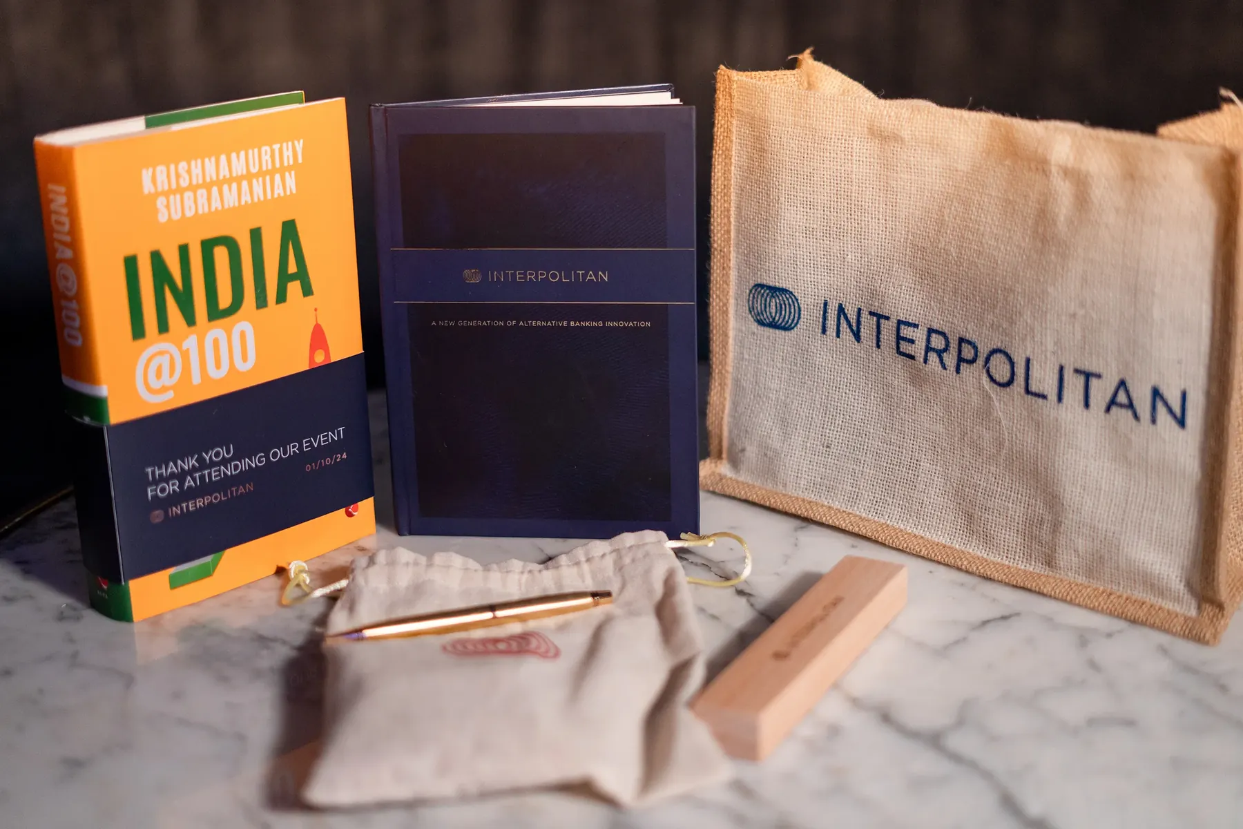 An Interpolitan-branded notebook, jute bag, pen and India@100 book on a marble surface.