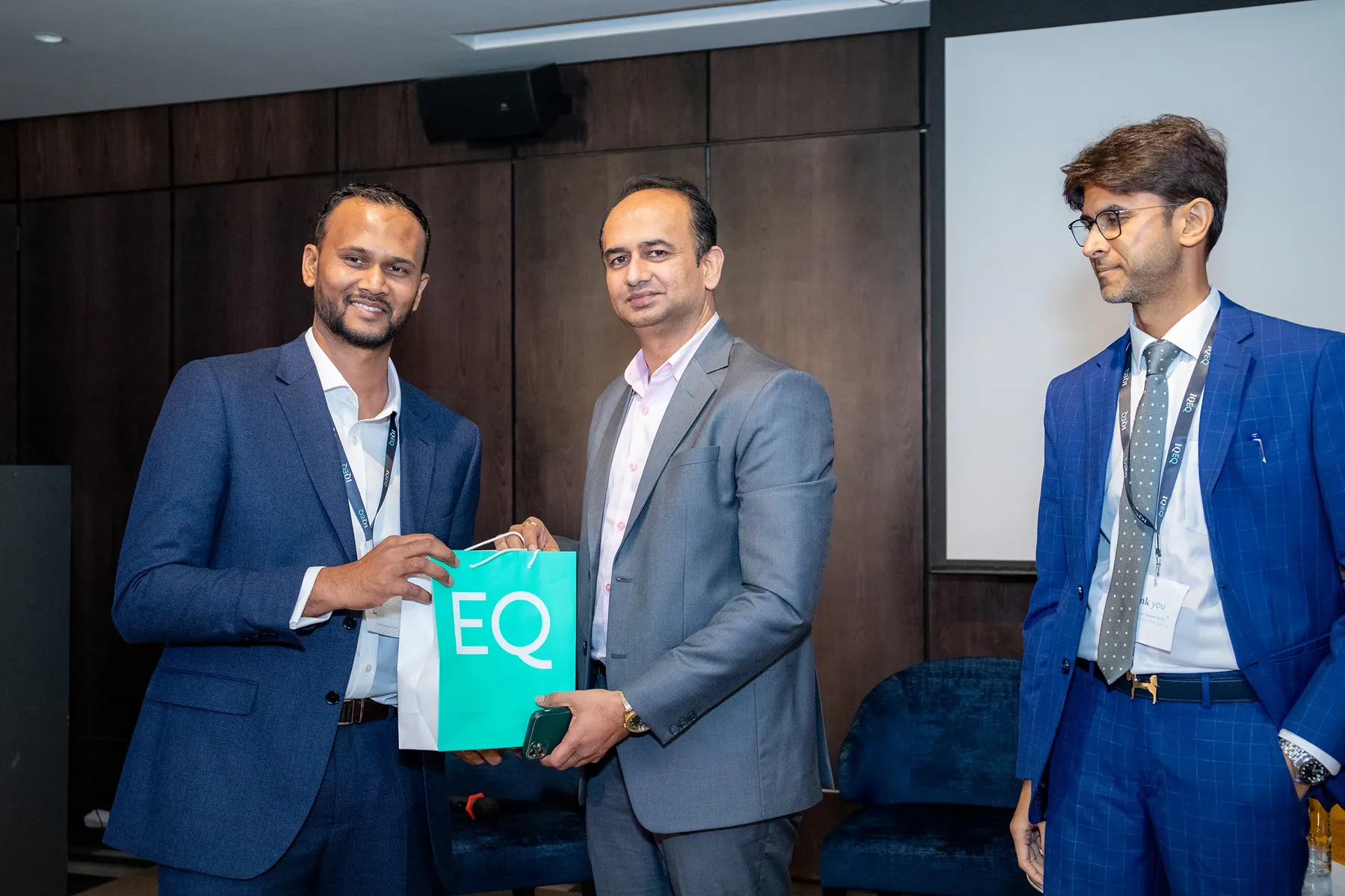 A man presents an IQ-EQ branded gift bag to another on a conference stage.
