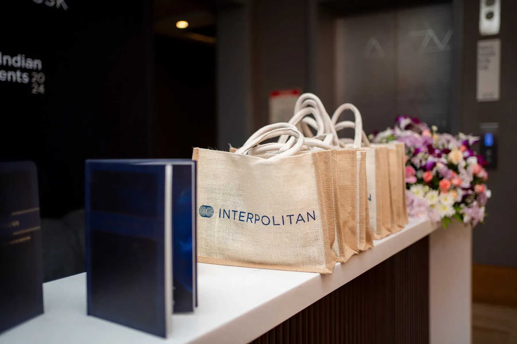 Interpolitan branded jute tote bags and gift boxes on a reception counter with flowers.