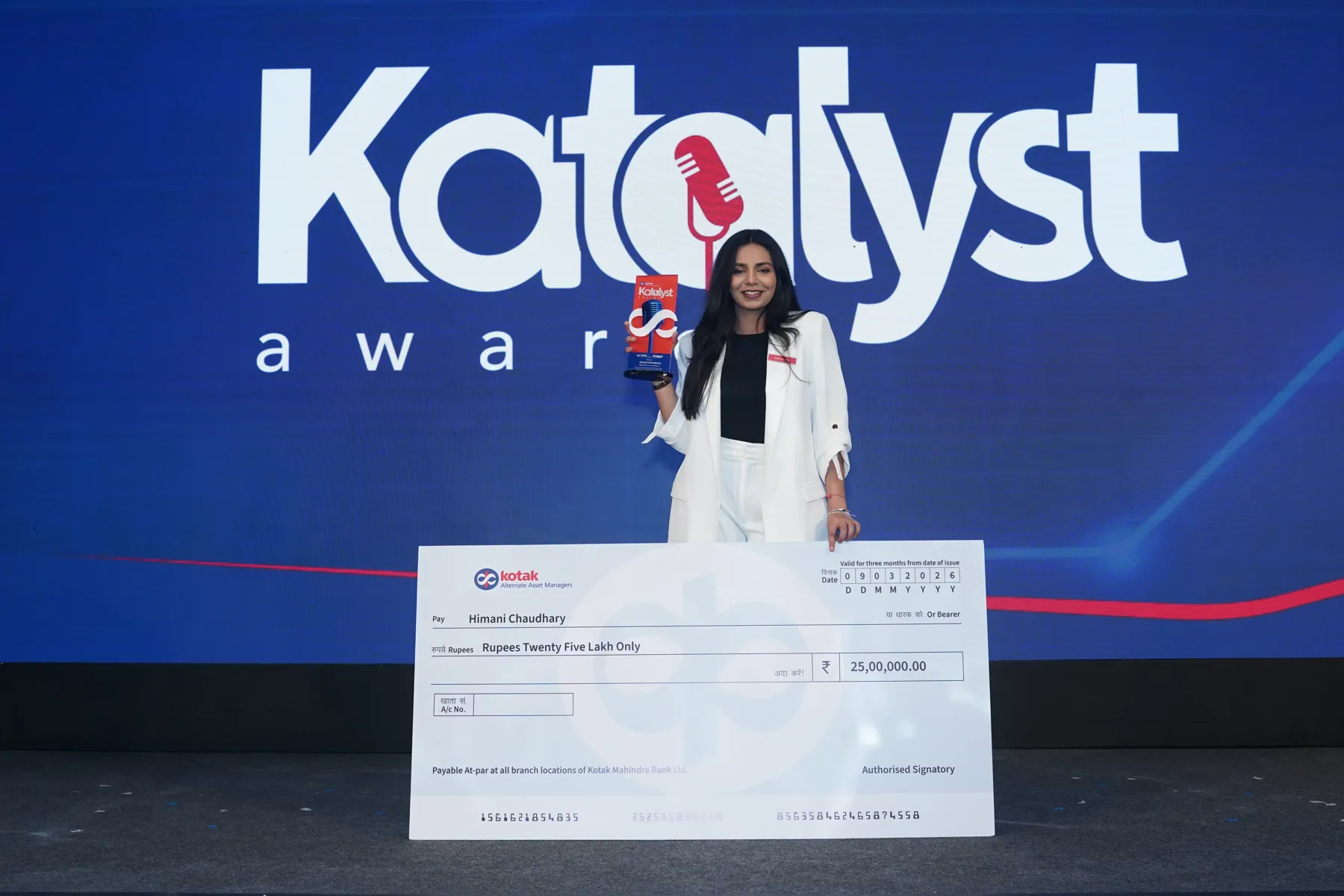 A woman in a white suit holds a trophy and oversized cheque before a Katalyst Awards LED backdrop.