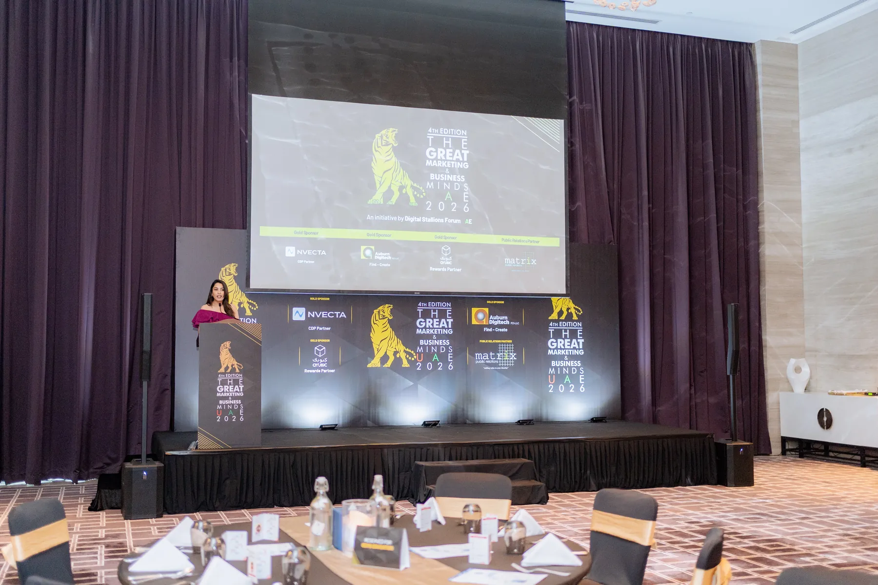 A speaker at a podium on stage before a large screen at Great Marketing and Business Minds UAE 2026.