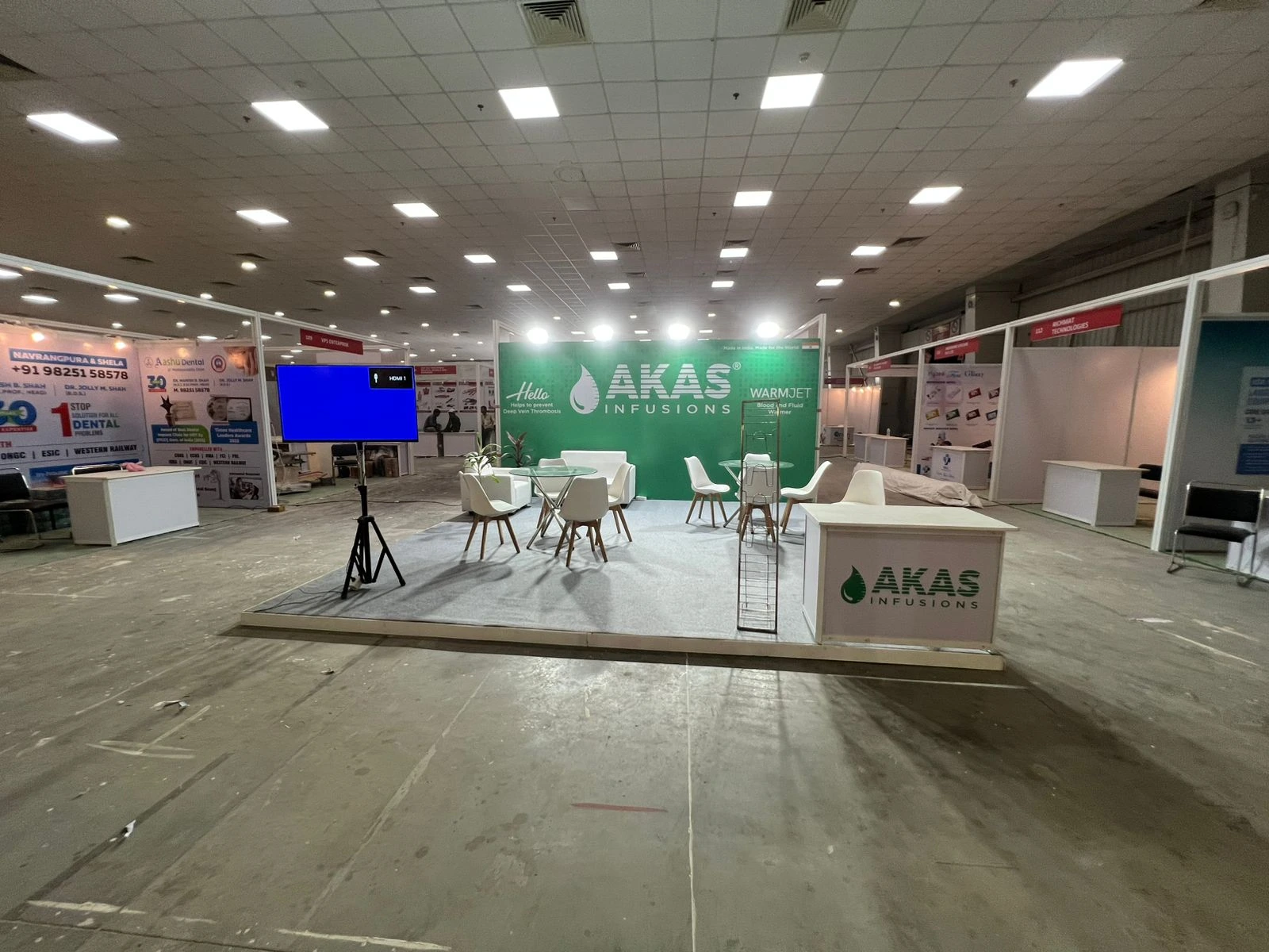 A wide view of the AKAS Infusions exhibition booth with seating and an LED screen in a convention hall.