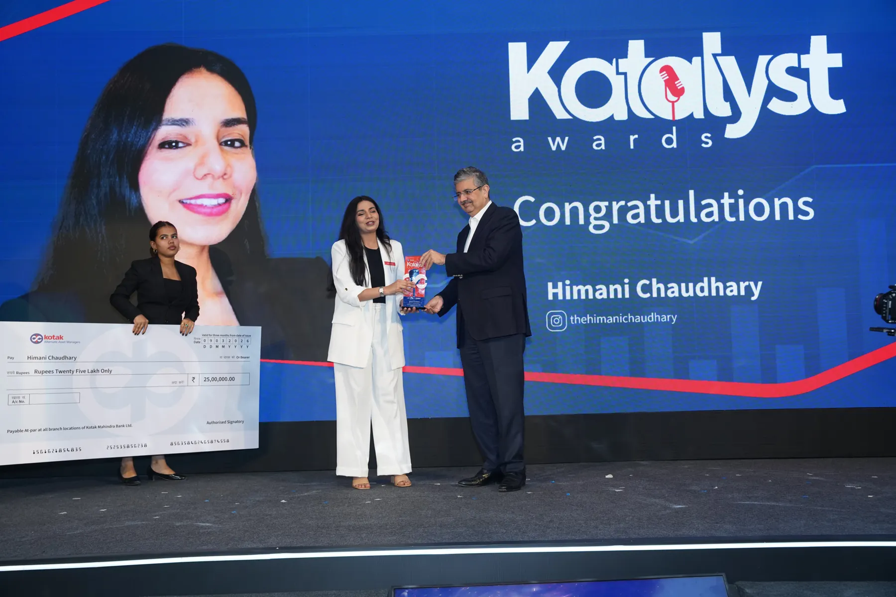 A woman in a white suit accepts a trophy beside an oversized cheque at the Katalyst Awards stage.