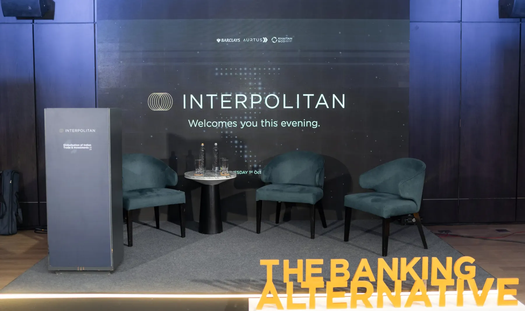 A stage with three teal armchairs and an Interpolitan backdrop set for The Banking Alternative.
